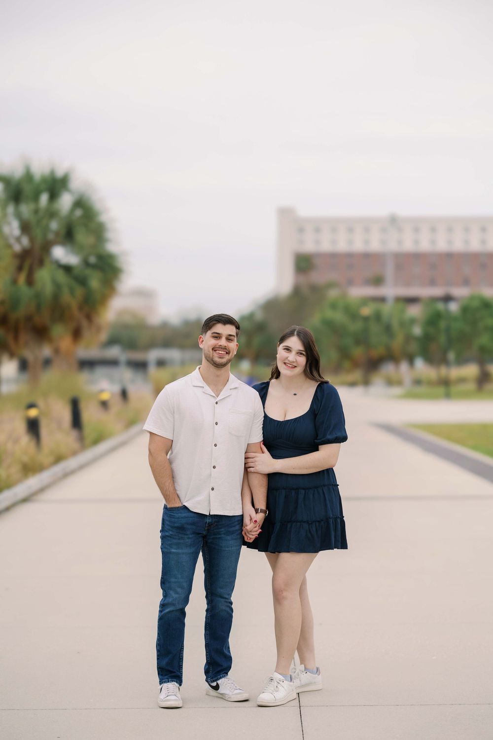 A couple stands smiling together on a concrete path with palm trees and buildings in the background.