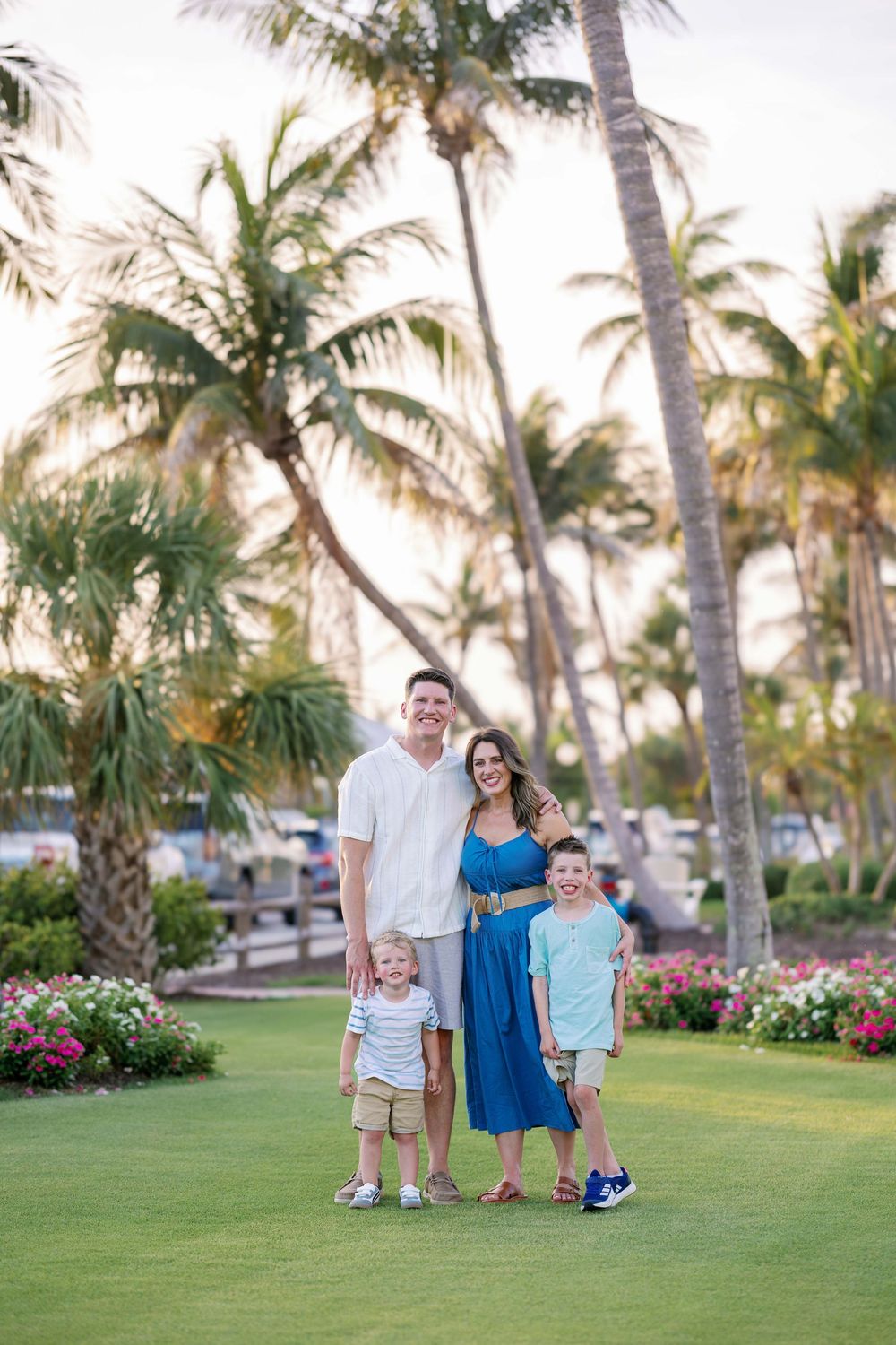 A family stands together on a manicured lawn with tropical palm trees and pink flowers in the background at sunset.