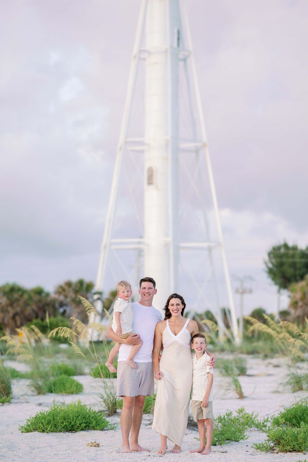 A family stands together on a sandy beach with white sailboats visible in the background on a cloudy day.