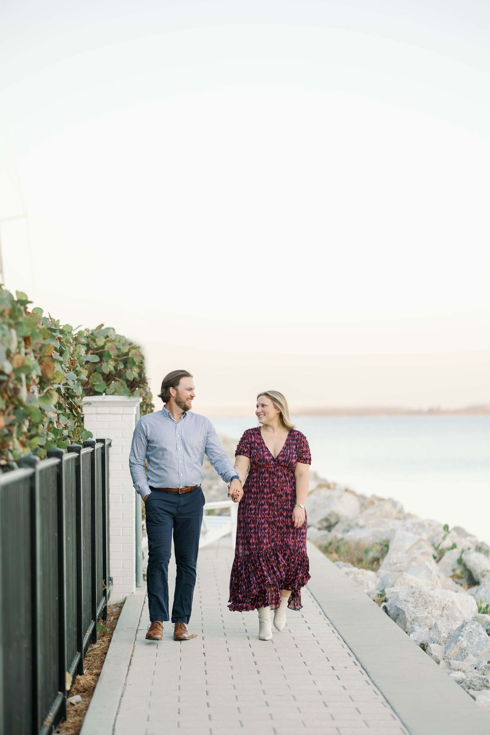 Couple holding hands walking on a coastal boardwalk at sunset, man in blue shirt and woman in floral dress.