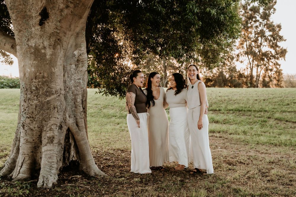 Four bridesmaids in white dresses standing together under a large tree in a sunlit green field.