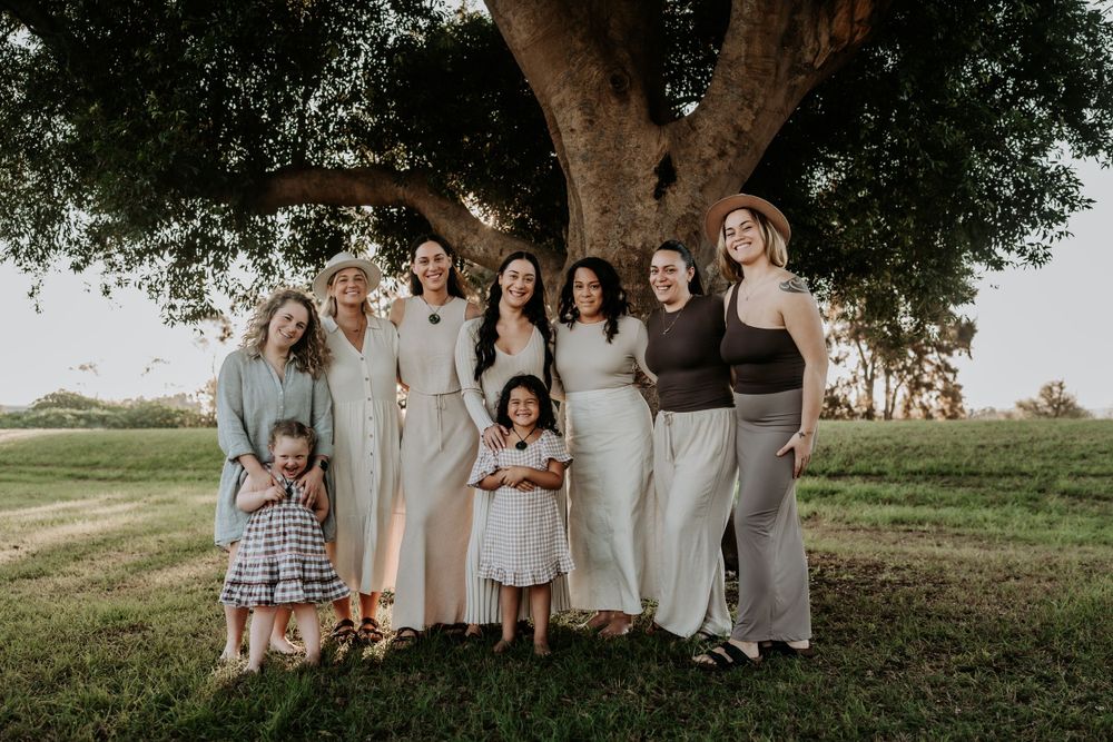 Group of eight women and two young girls smiling together under a large tree on a green lawn.