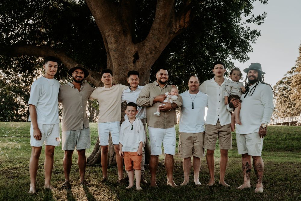 Group of nine men and boys posing together under a large tree on a sunny green lawn.