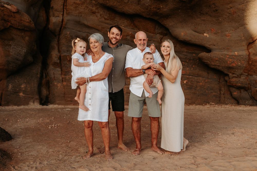 Multi-generational family smiling together at a red rock beach location holding two babies.