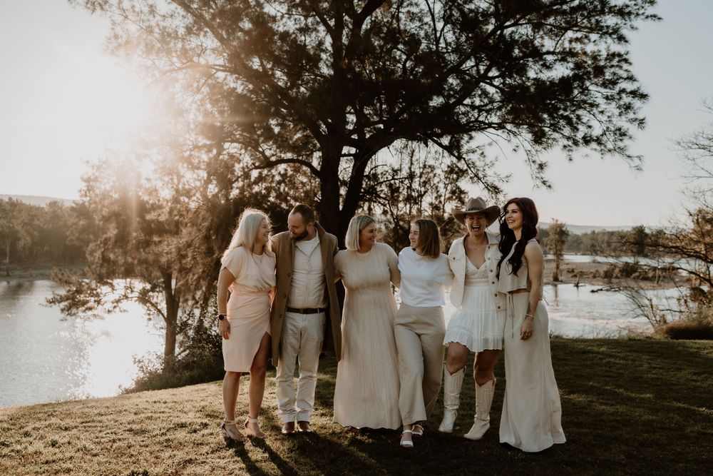 Group of women in white and neutral outfits laughing together by sunlit lakeside.