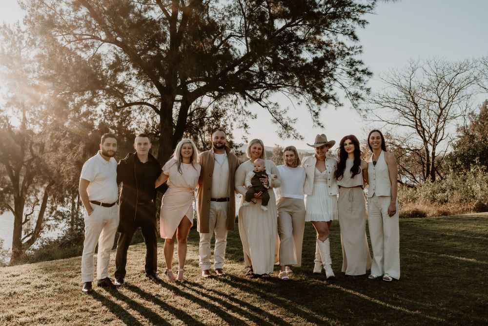 Large family group portrait outdoors at golden hour near a tree and lake, dressed in elegant white and neutral tones.