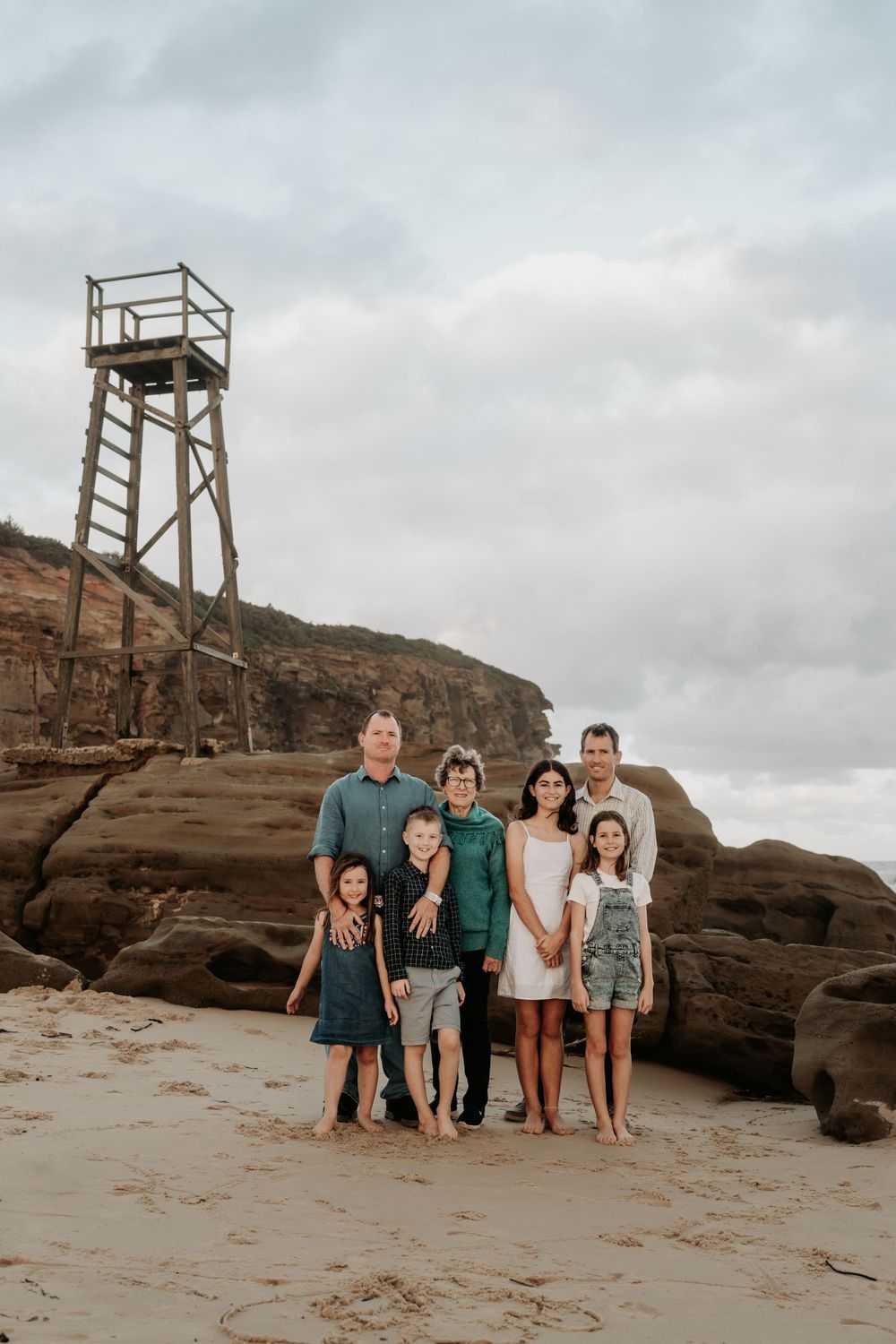 Family of seven posing on a sandy beach with dramatic rock cliffs and a lifeguard tower in the background.