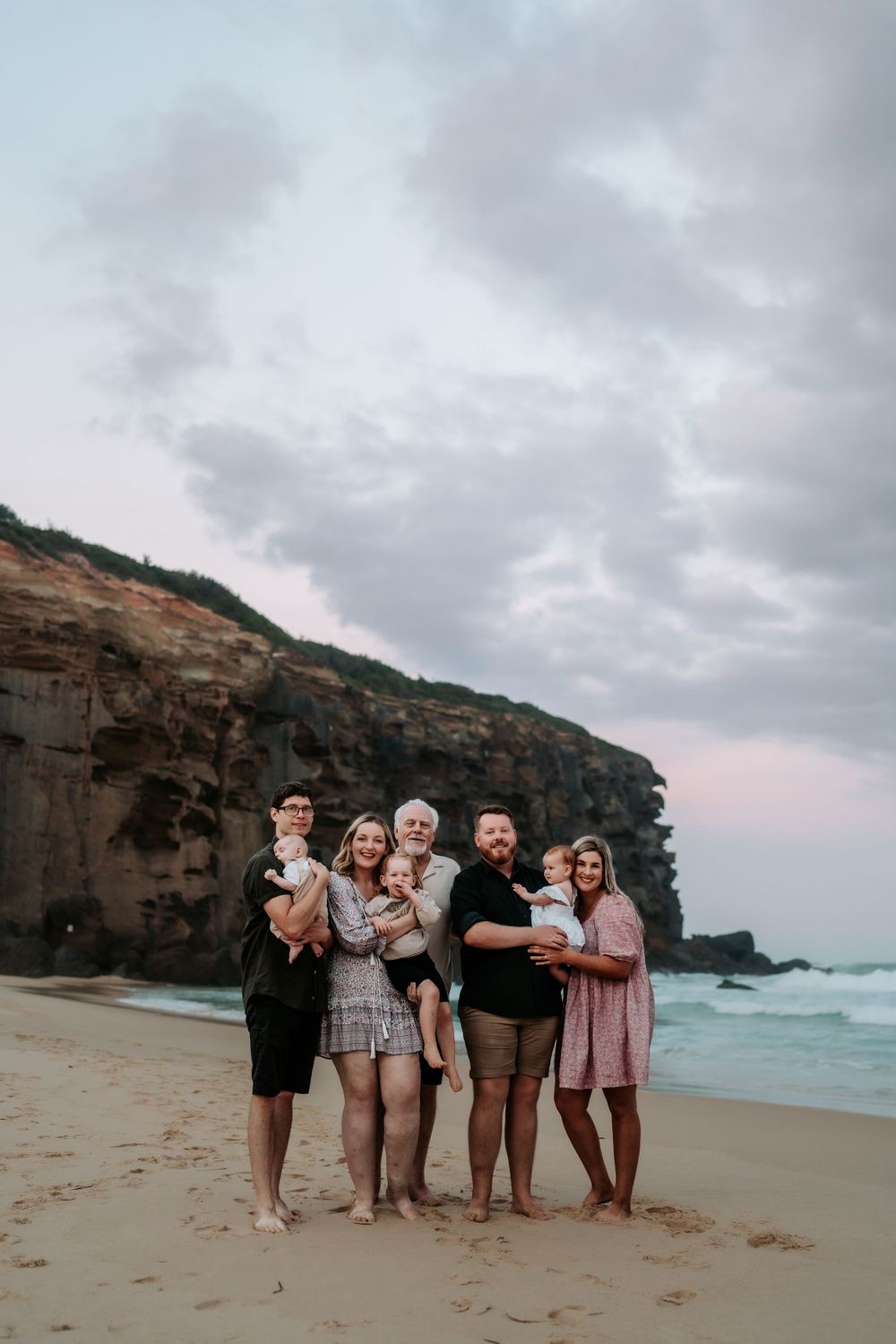 Multi-generational family of seven smiling together on a sandy beach at sunset with rocky cliffs behind.
