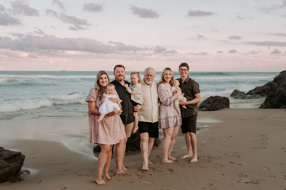 Multi-generational beach family portrait at sunset with ocean waves and pastel sky in the background.