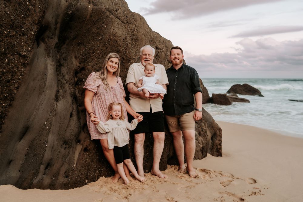 Three-generation family of five posing against a large rock on a beach at dusk with ocean waves behind.
