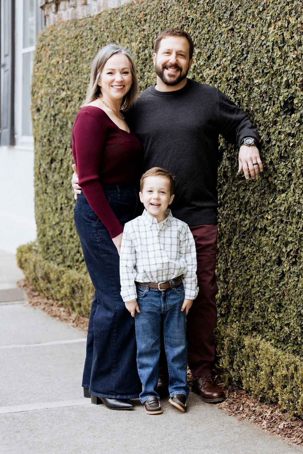 A family poses together against a green hedge wall in coordinated casual outfits for outdoor portraits.