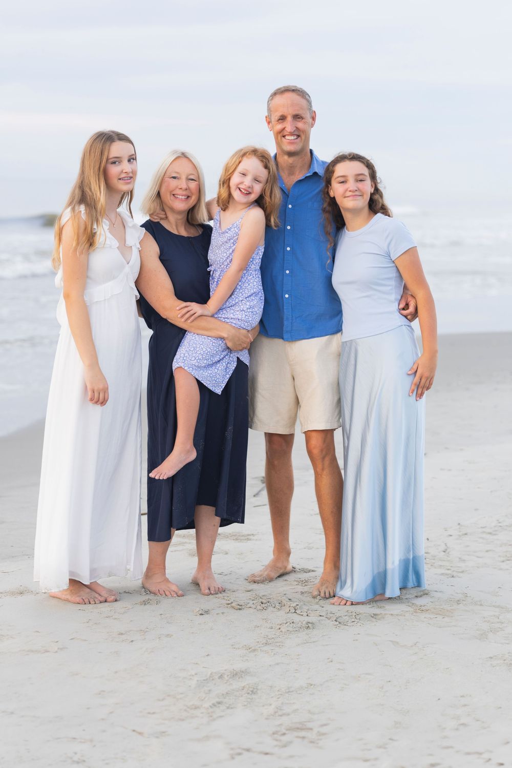 Family group wearing coordinated blue and white outfits poses together on sunny beach at sunset.