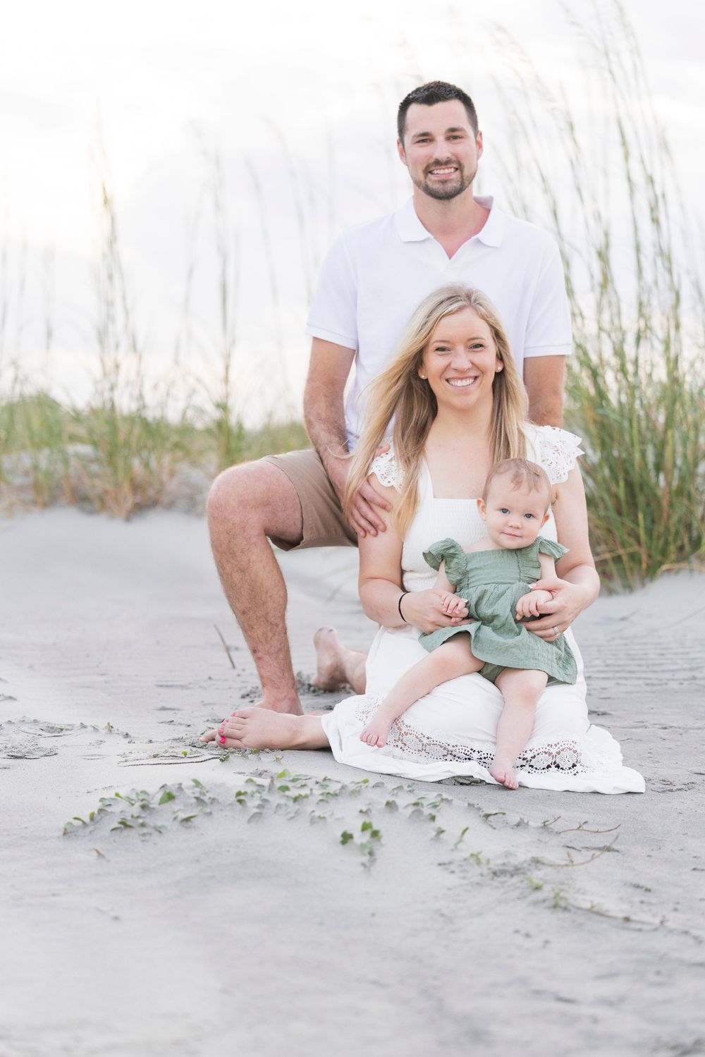 A family enjoys a casual photo session on a sandy beach with sea oats in the background.