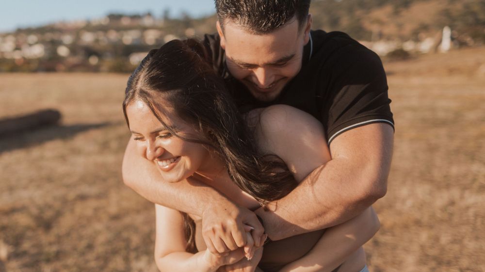 Man hugging a smiling woman outdoors with dry grassland and distant buildings in the background.