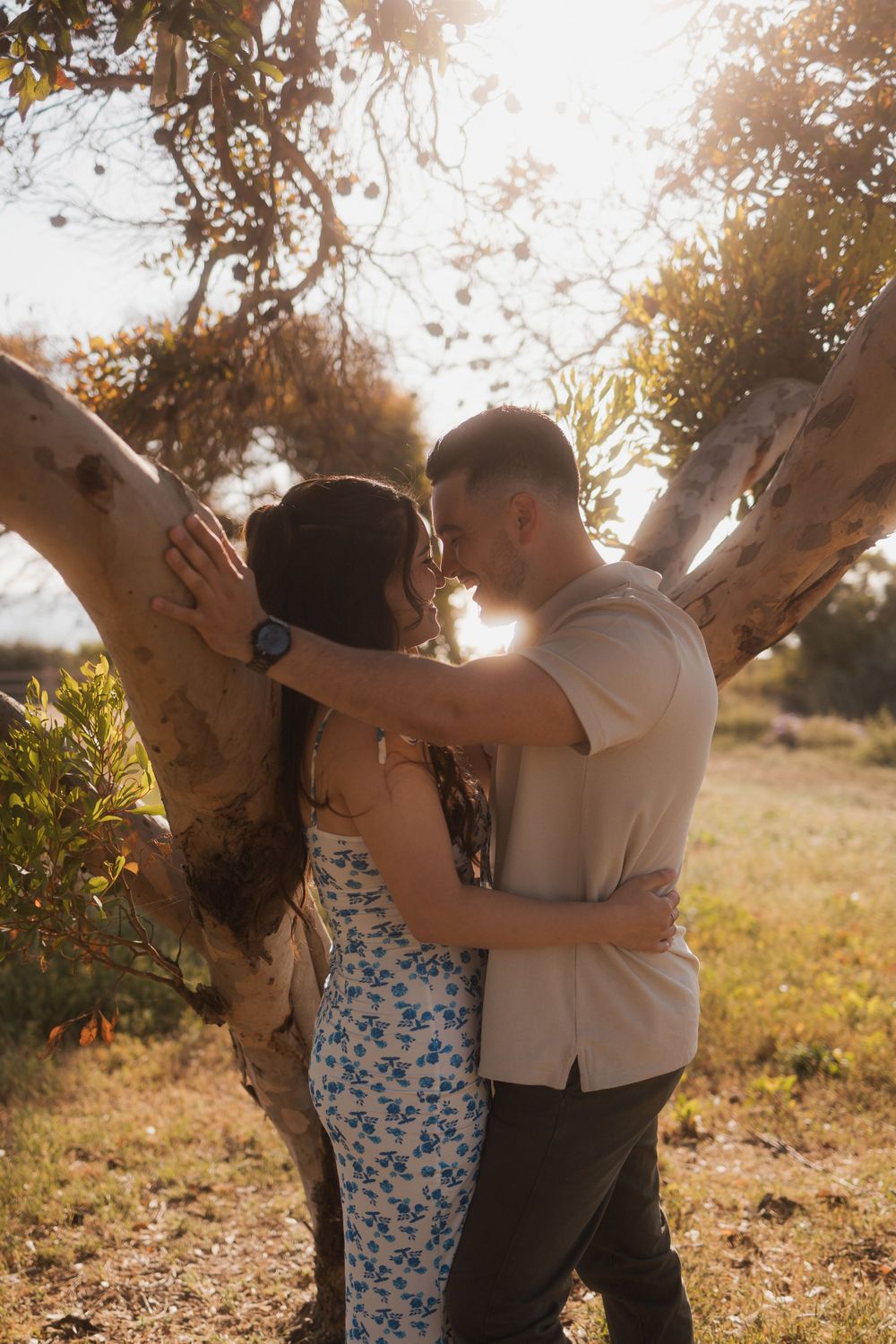 A couple standing closely under a tree with their foreheads touching, illuminated by setting sun.