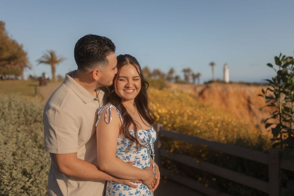 Man hugging woman outdoors with yellow flowers and lighthouse in the background.