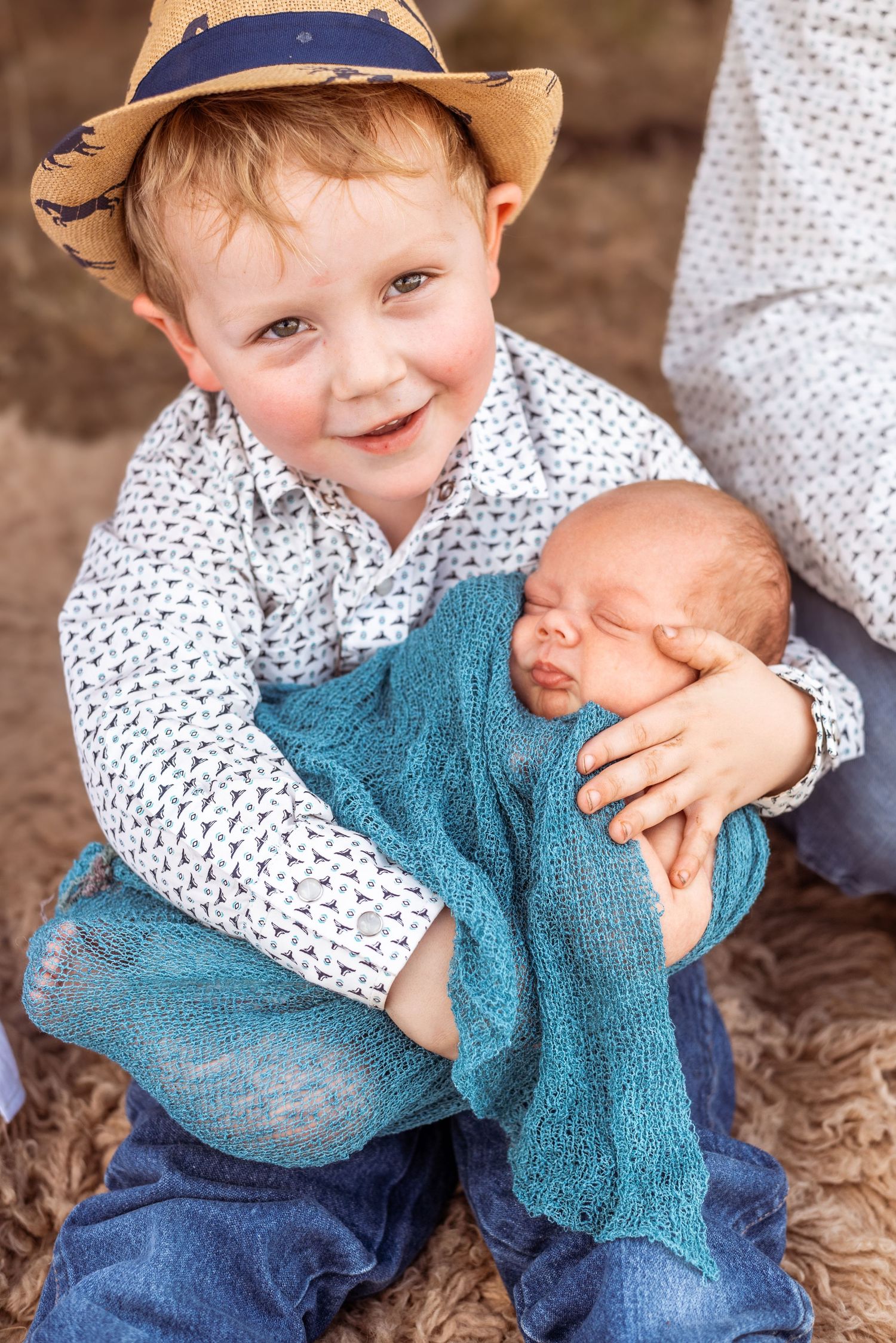 Young child in a hat holds a sleeping newborn wrapped in a teal blanket with a joyful expression.