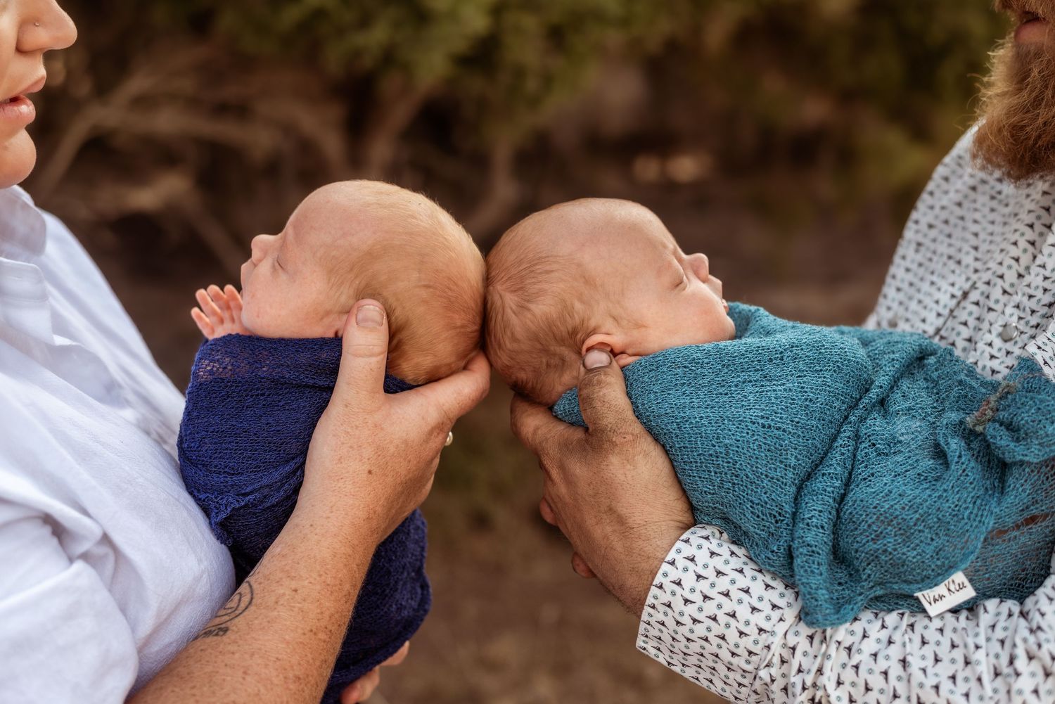 Two adults holding newborn babies wrapped in blue blankets during outdoor photoshoot.