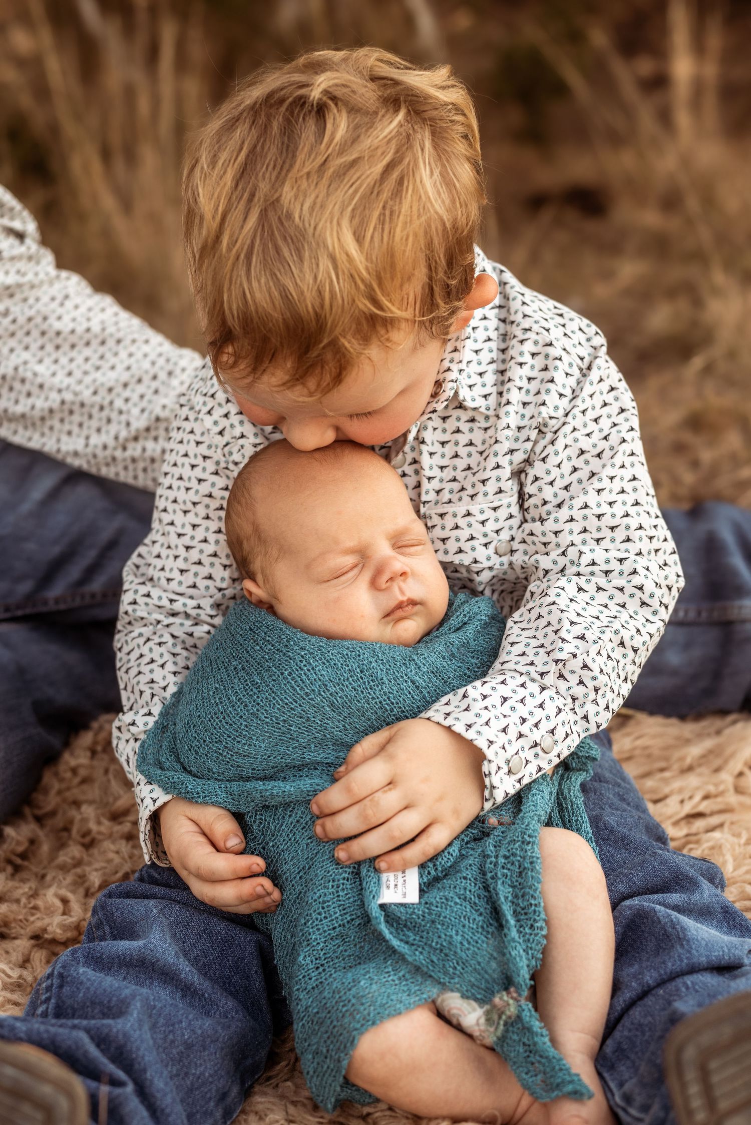Sweet sibling moment as child in patterned shirt gives gentle kiss to sleeping baby in teal wrap.