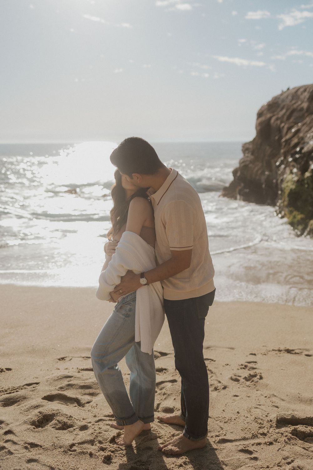 Couple embracing on a sandy beach with ocean and rocky outcrop in the background.