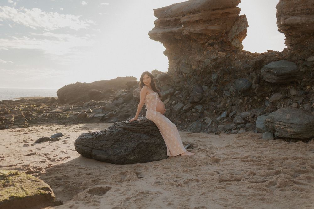 A pregnant woman in a pale, strapless dress sits on a large rock on a sandy beach with rocky formations