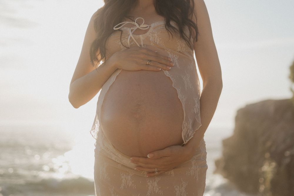 Close-up of a pregnant woman wearing a beige lace dress, cradling her belly with both hands.