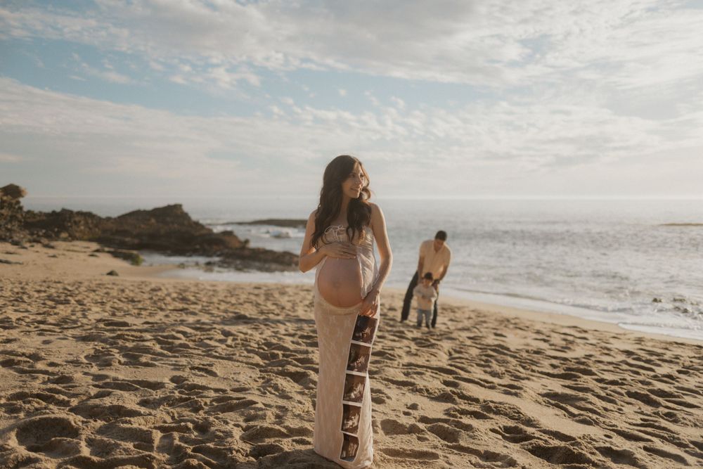 Pregnant woman in a lace dress stands on a beach with ultrasound photos on her dress, while a man and child interact