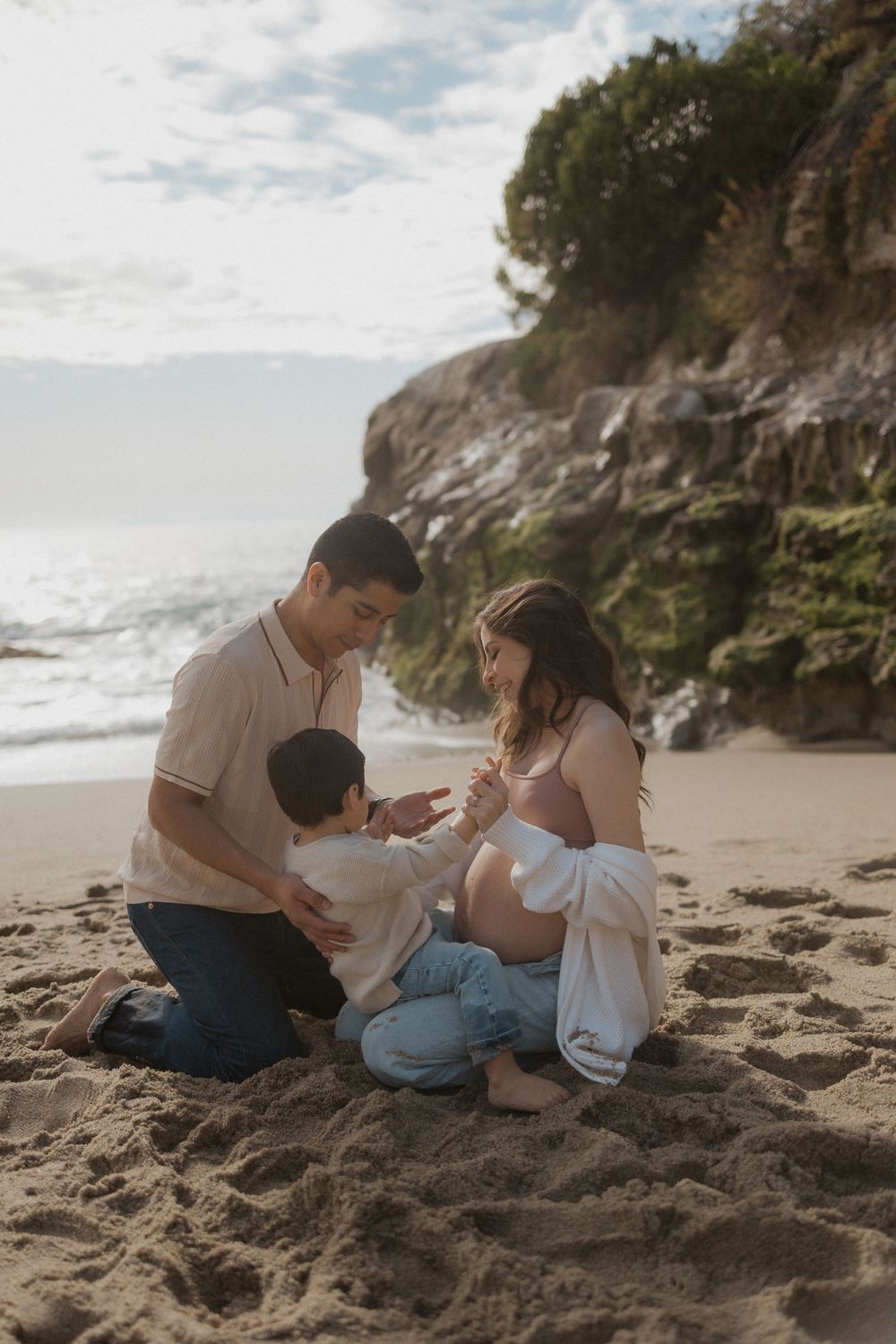 Man, pregnant woman, and child holding hands on a beach with the ocean and cliffs in the background.