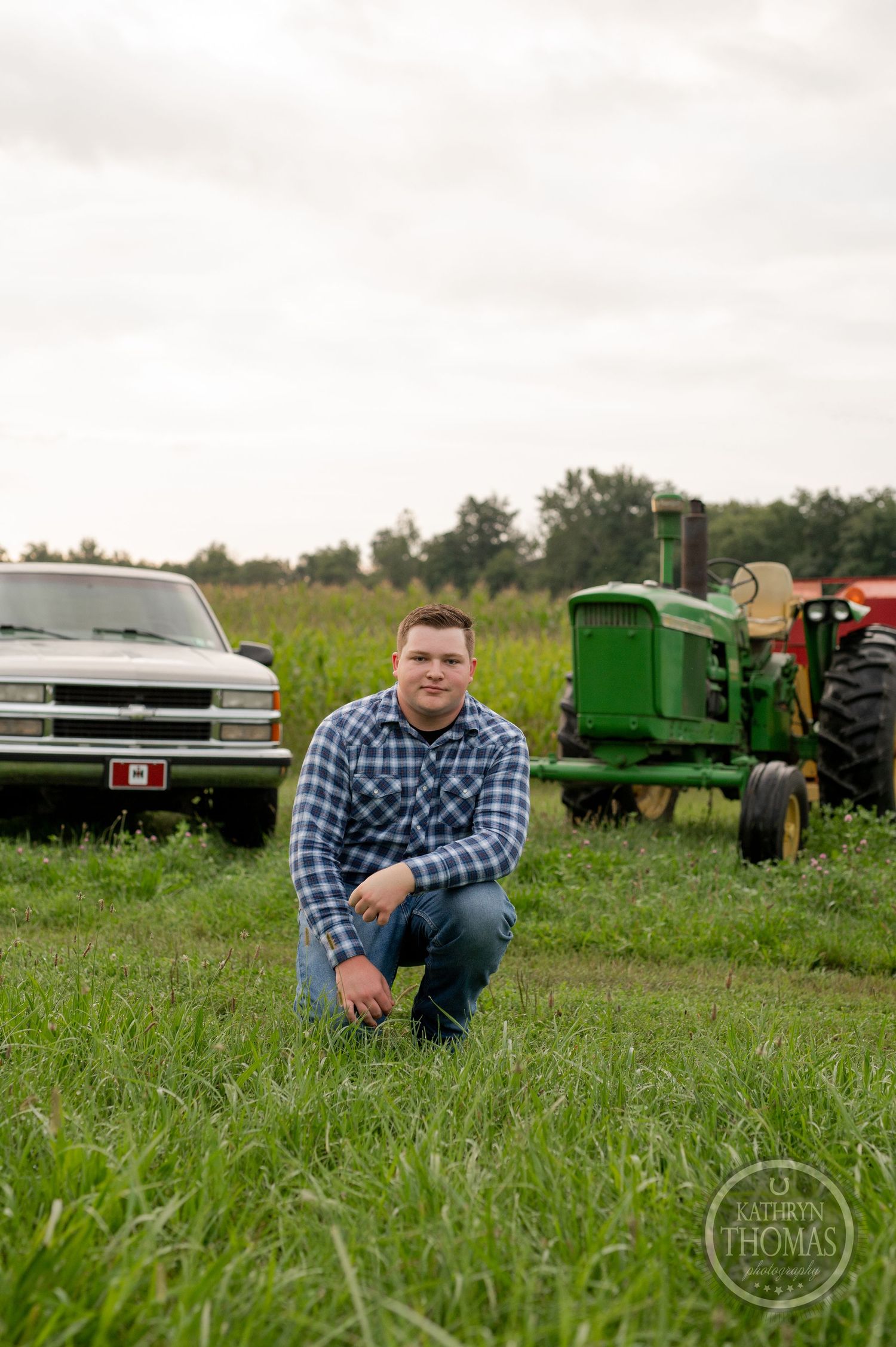 A person kneels in front of a silver Chevy pickup truck and classic green tractor in a grassy field.
