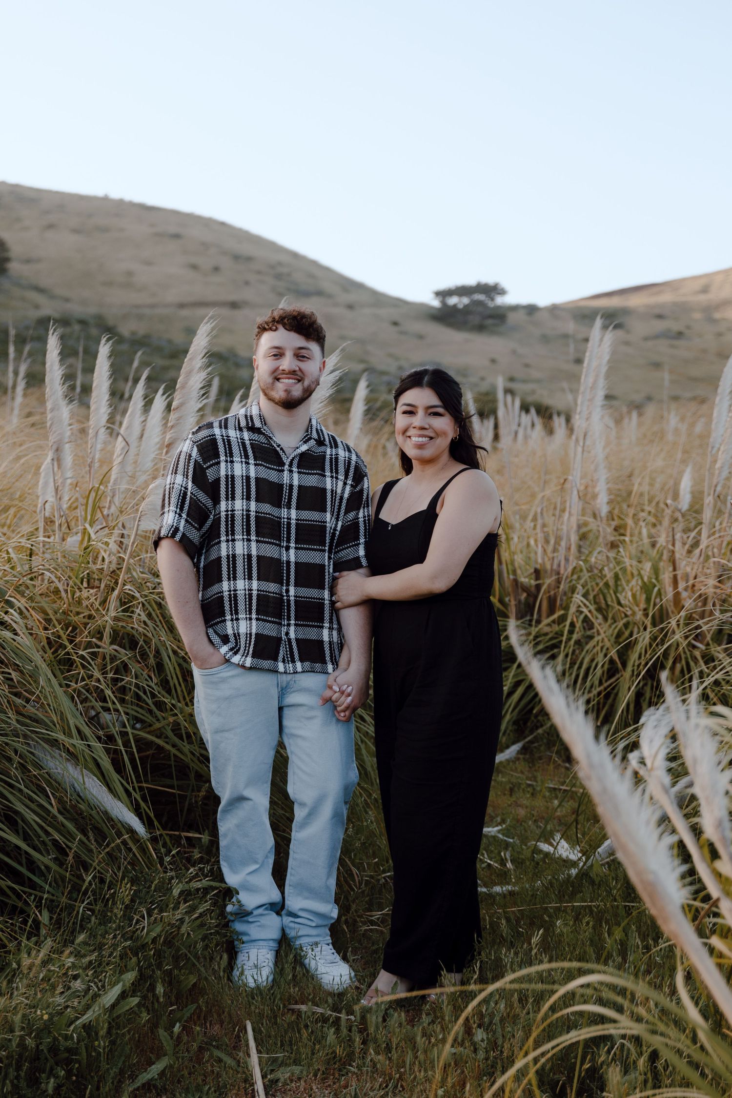 Couple standing together in tall pampas grass field with rolling hills in background.