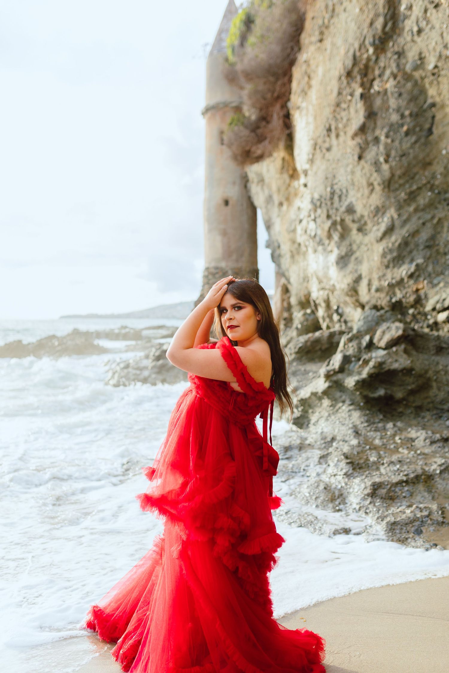A woman in a flowing red gown poses dramatically against coastal cliffs and ocean waves.