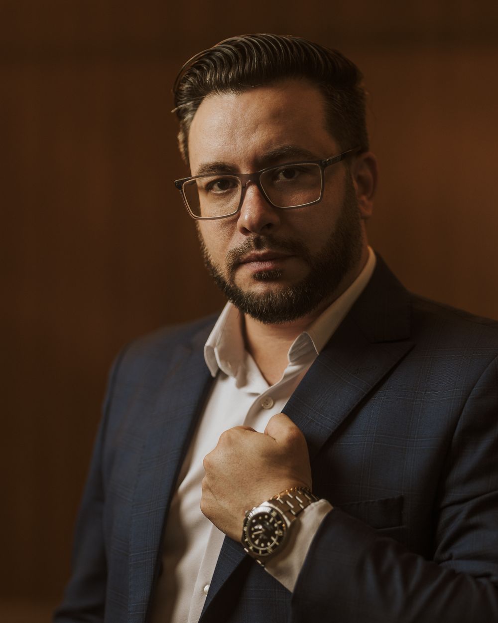 Professional business portrait of a person in a dark suit wearing glasses and a luxury watch against a brown backdrop.