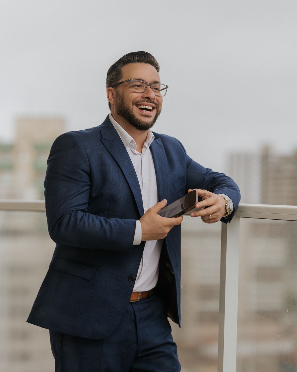 Business professional in navy suit smiling while checking phone on an outdoor balcony.