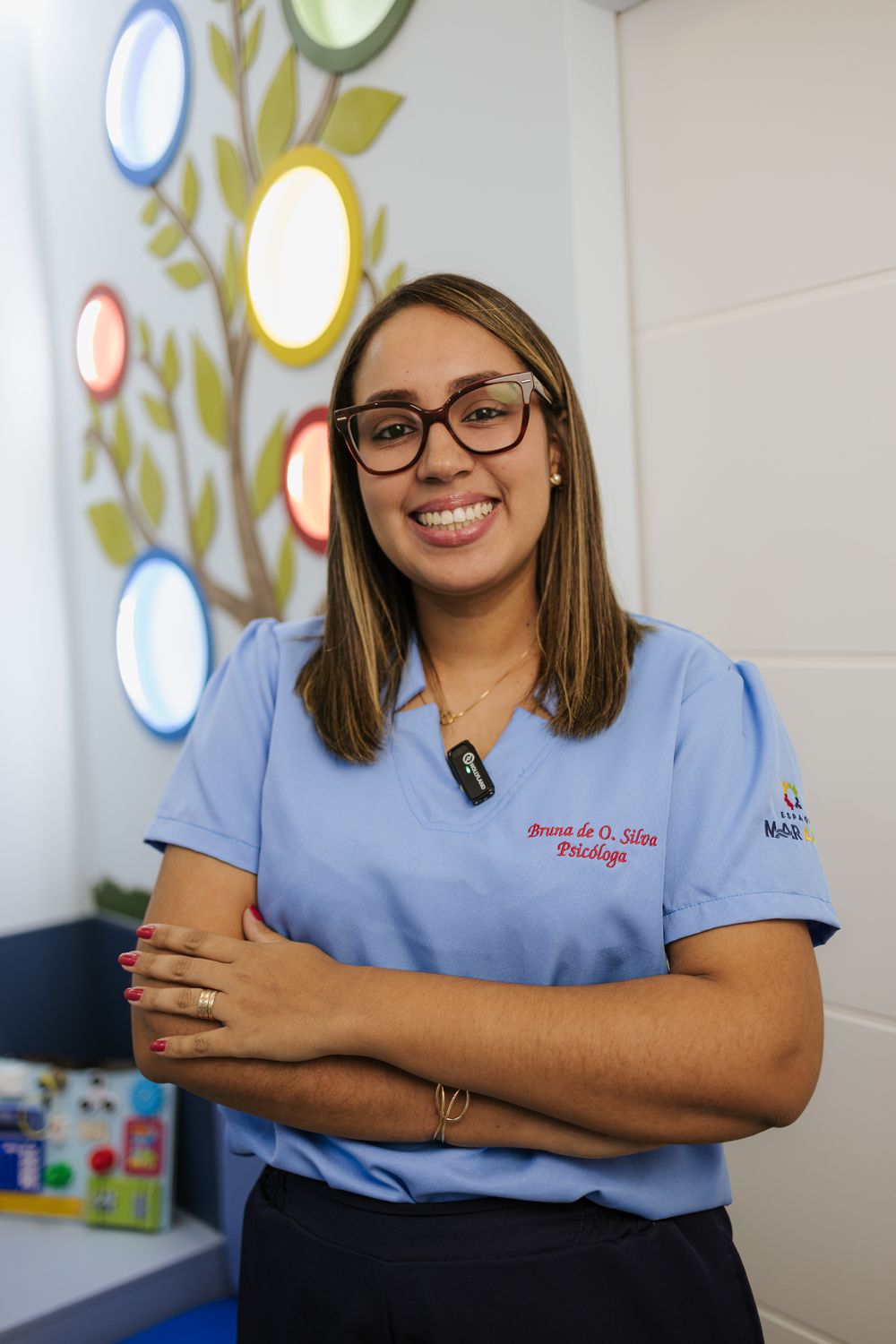 Healthcare worker in blue scrubs poses professionally against wall with decorative tree mural.