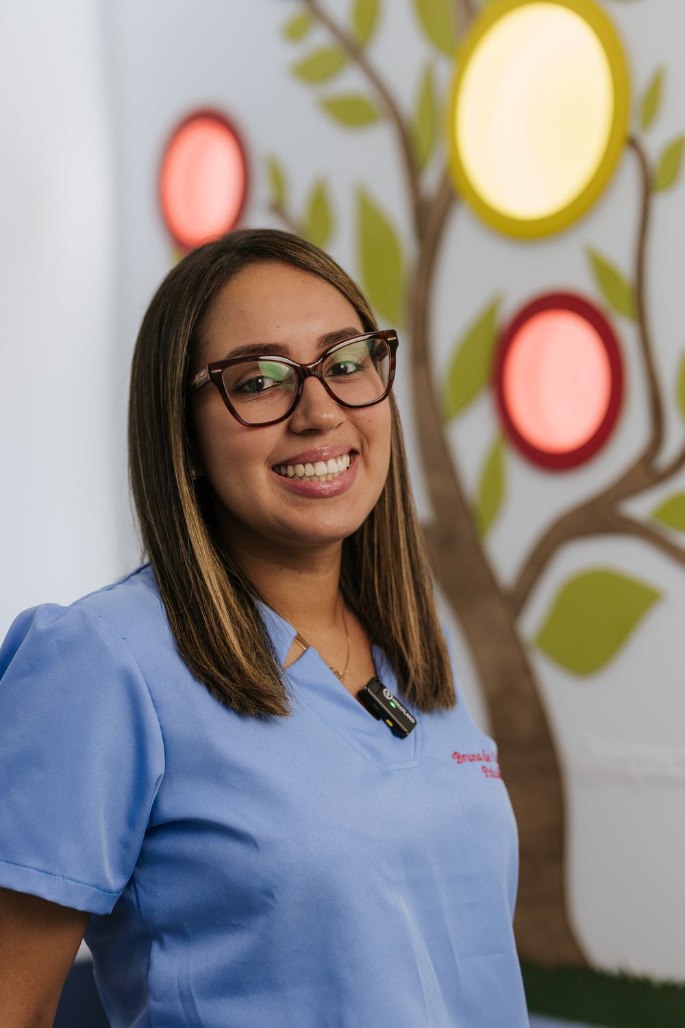Medical professional in scrubs smiles warmly against colorful wall art background.