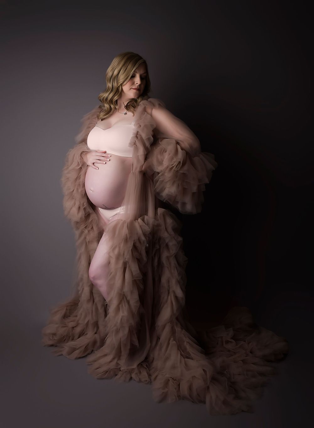 Maternity image on grey backdrop with tulle dress. Looking to the side and rested hand on top of the bump. Dark and shadowy.