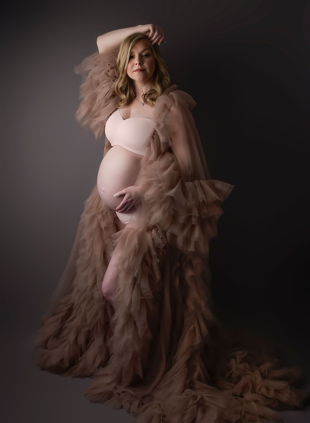 Maternity image on grey backdrop with tulle dress. Looking to the side and rested hand on top of head. Dark and shadowy.