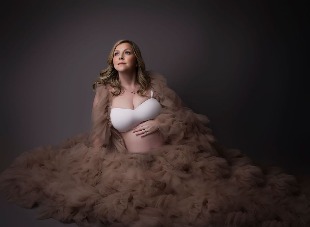 Maternity image looking off to the side with dark shadowy light. Hand is rested on the bump in a tulle beige dress.