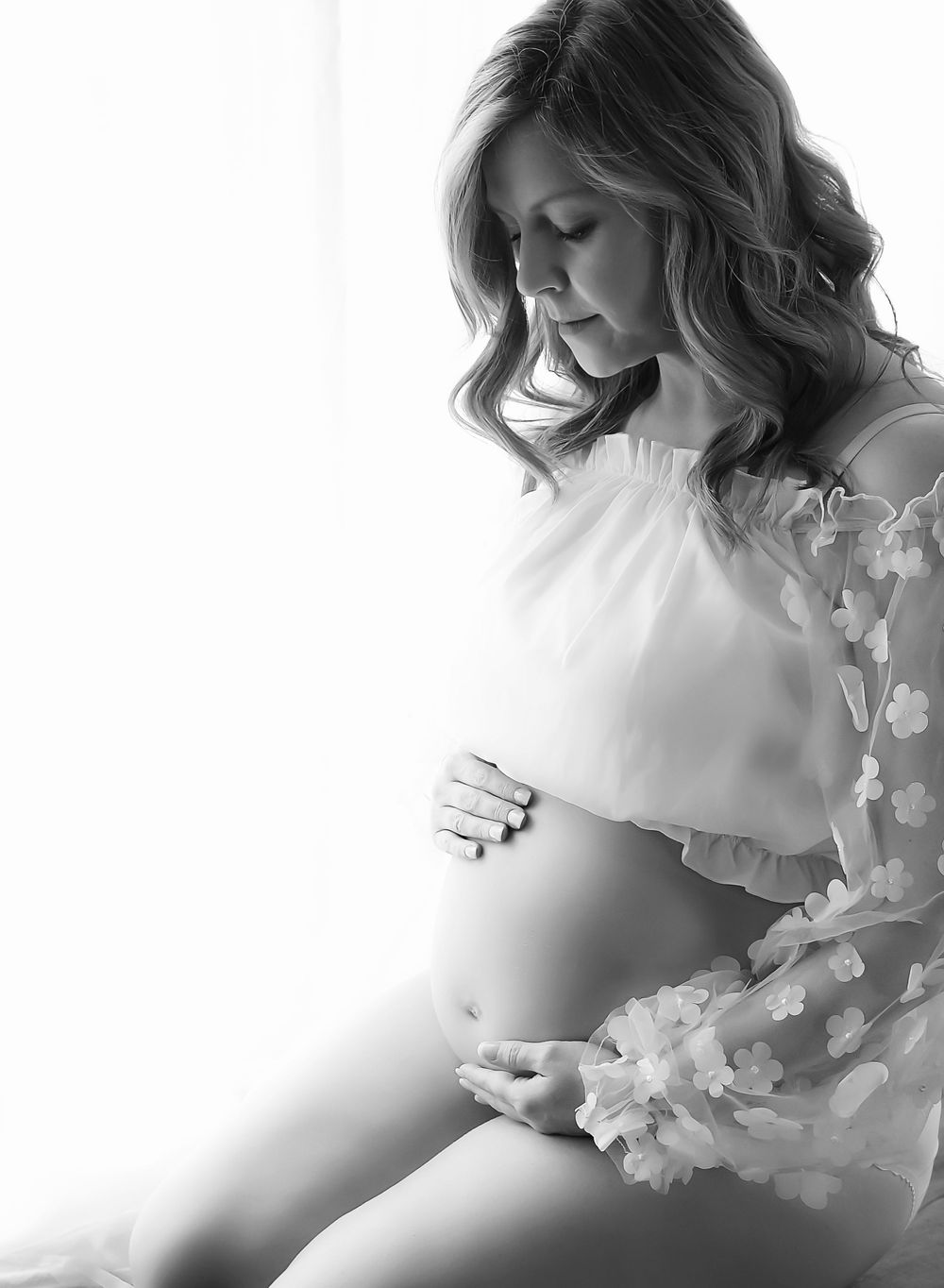 Black and white maternity image. Backlit window scene. Mother looking down at their bump and hands rested.