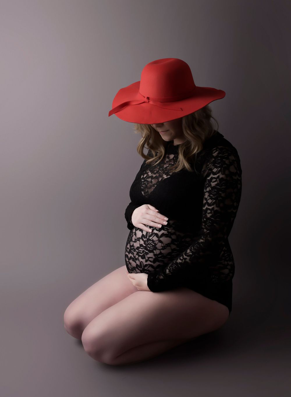 Maternity image, mum knelt on the floor in a red hat clutching her baby bump. Grey backdrop with dark shadows.