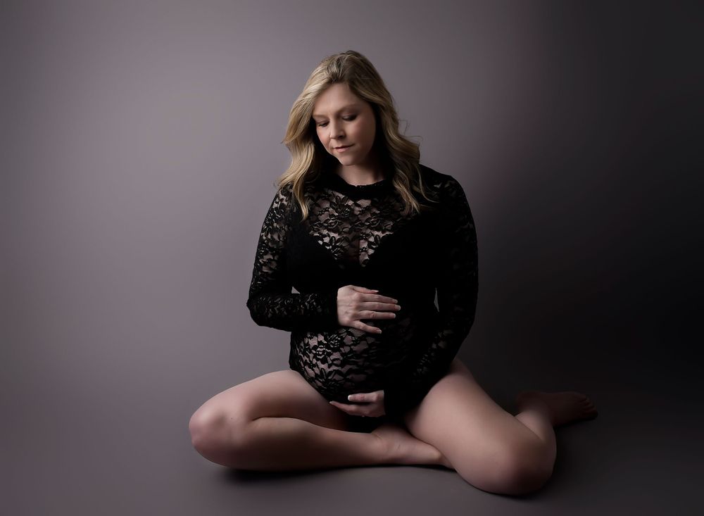 Maternity image on grey shadowy backdrop. Model in a black lacy body suit. looking at her bump and clutching it lovingly.