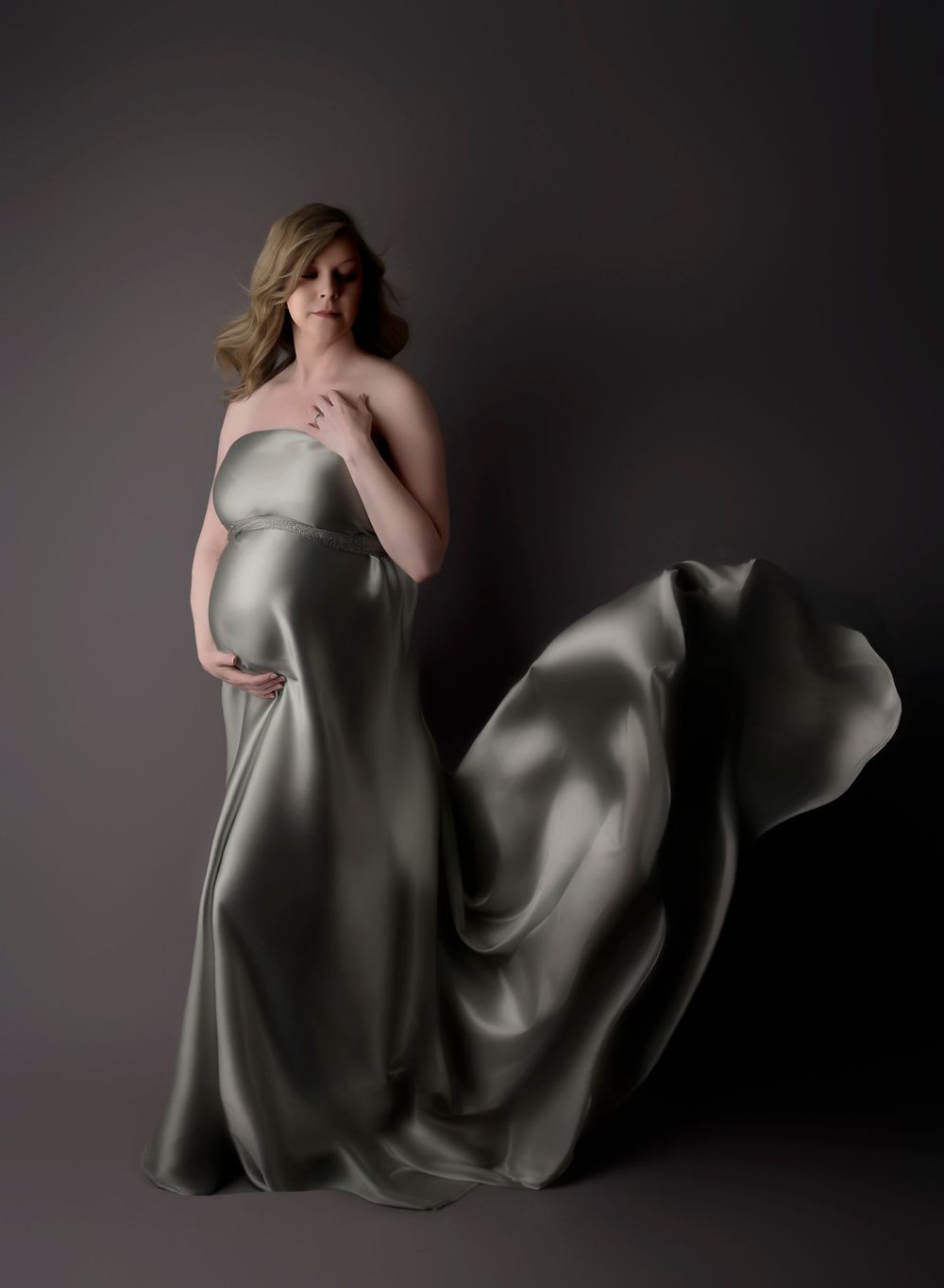 Maternity image with silk grey fabric. On a grey shadowy backdrop with the fabric cascading behind as her hand rests at her chest.