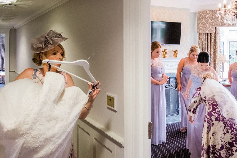 Wedding dress being carried through doorway with bridesmaids in lavender dresses visible in background.