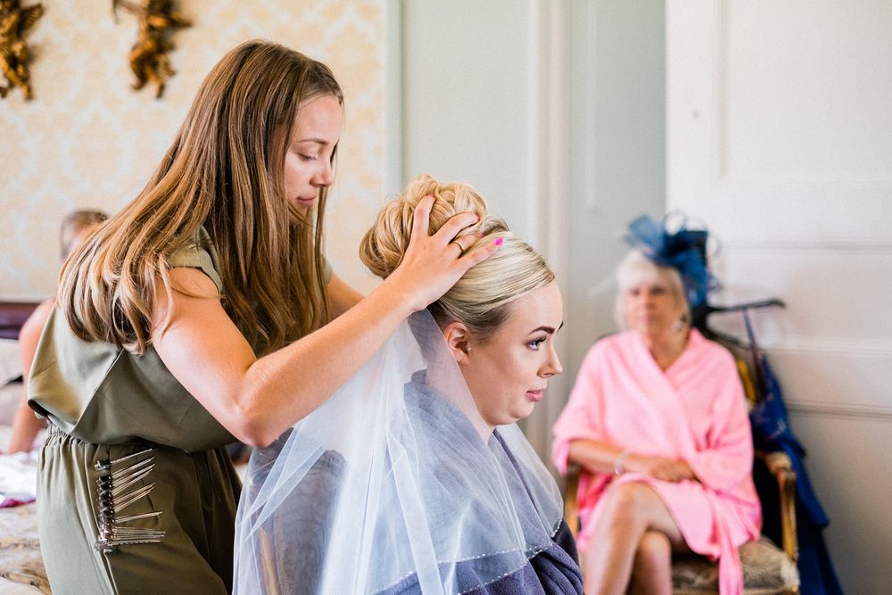 Hair stylist works on wedding day preparations in an elegant getting ready room with pink robed figure in background.