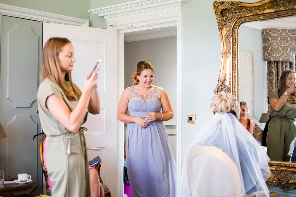 Bridesmaids in lavender dresses prepare for wedding day in elegant room with ornate mirror.
