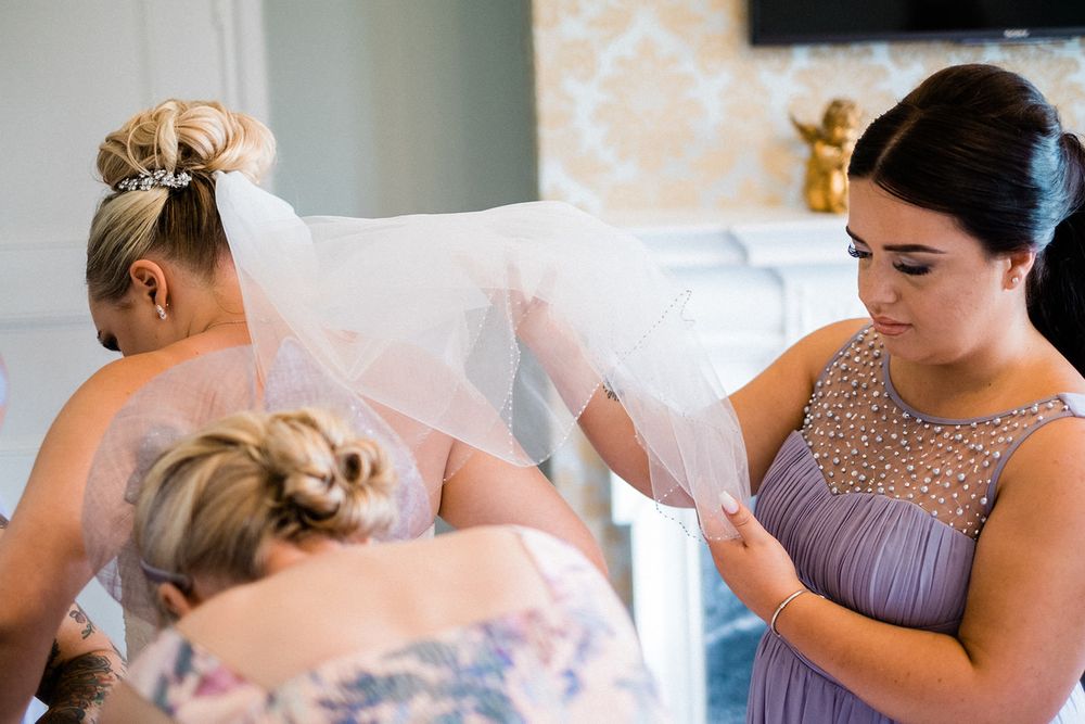 Bridesmaids in purple dresses help adjust wedding dress and veil during preparations.