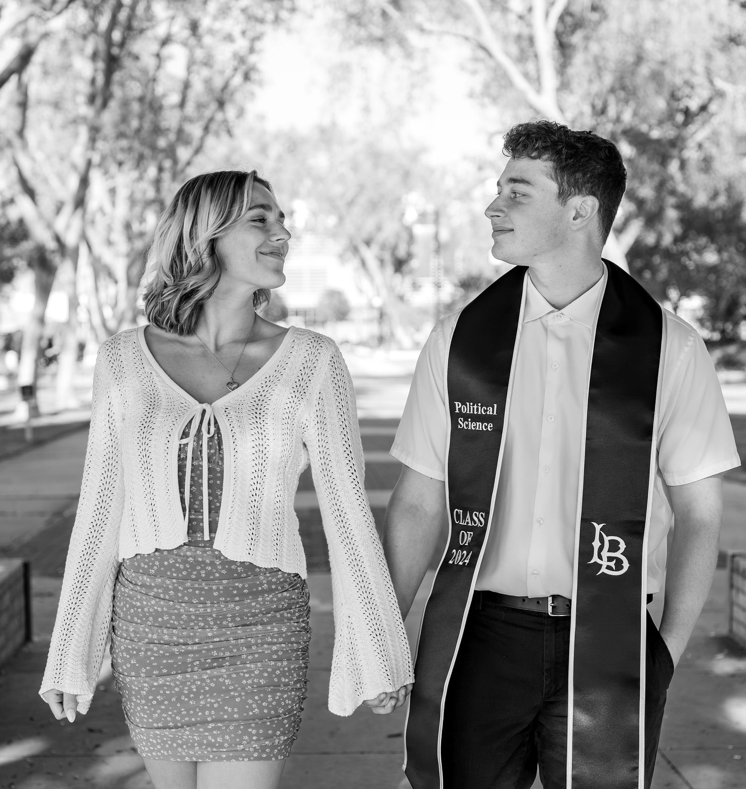 A couple in formal attire poses outdoors together, with one wearing a graduation stole and the other in a white knit top.