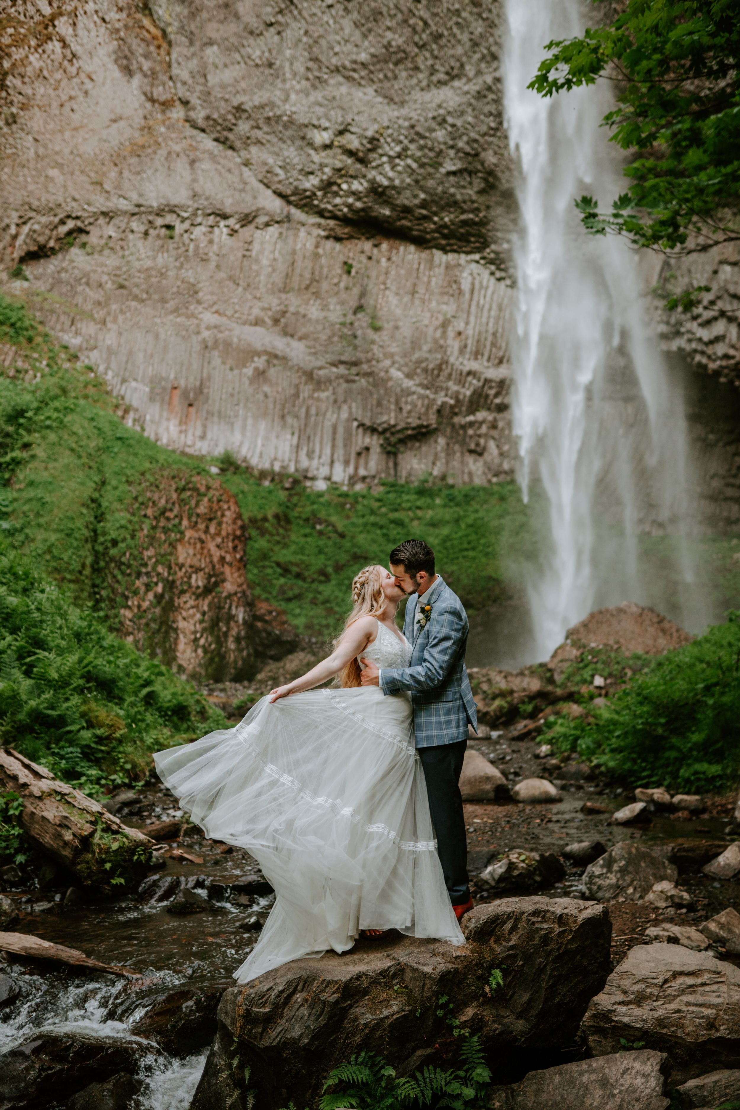 A couple embraces on rocky terrain with a majestic waterfall cascading in the background.