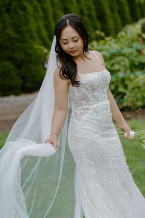 A bride in a beaded lace wedding gown holds her flowing veil in an outdoor garden setting.