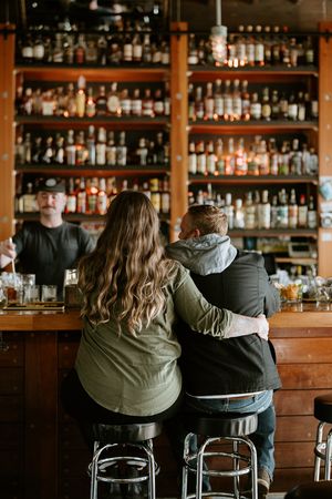 Two people sit side by side at wooden bar stools in front of a fully-stocked liquor shelf at a cozy bar.